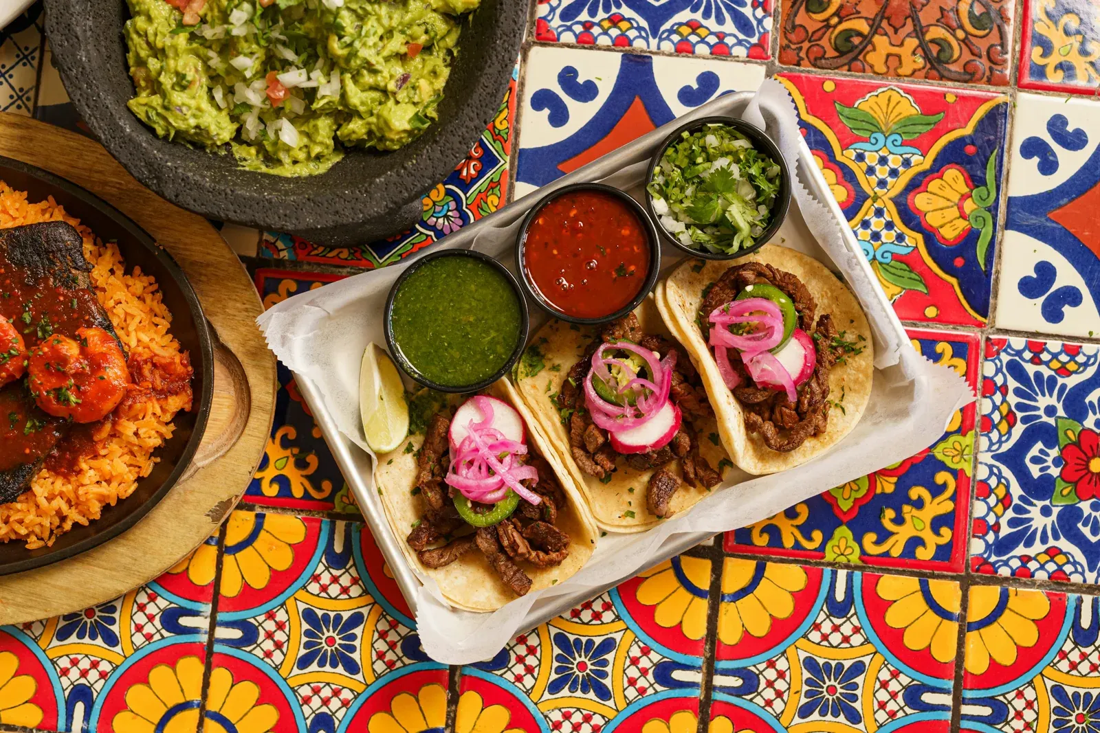 Tacos with radishes, pickled onions, salsa and guacamole on a colorful tiled table